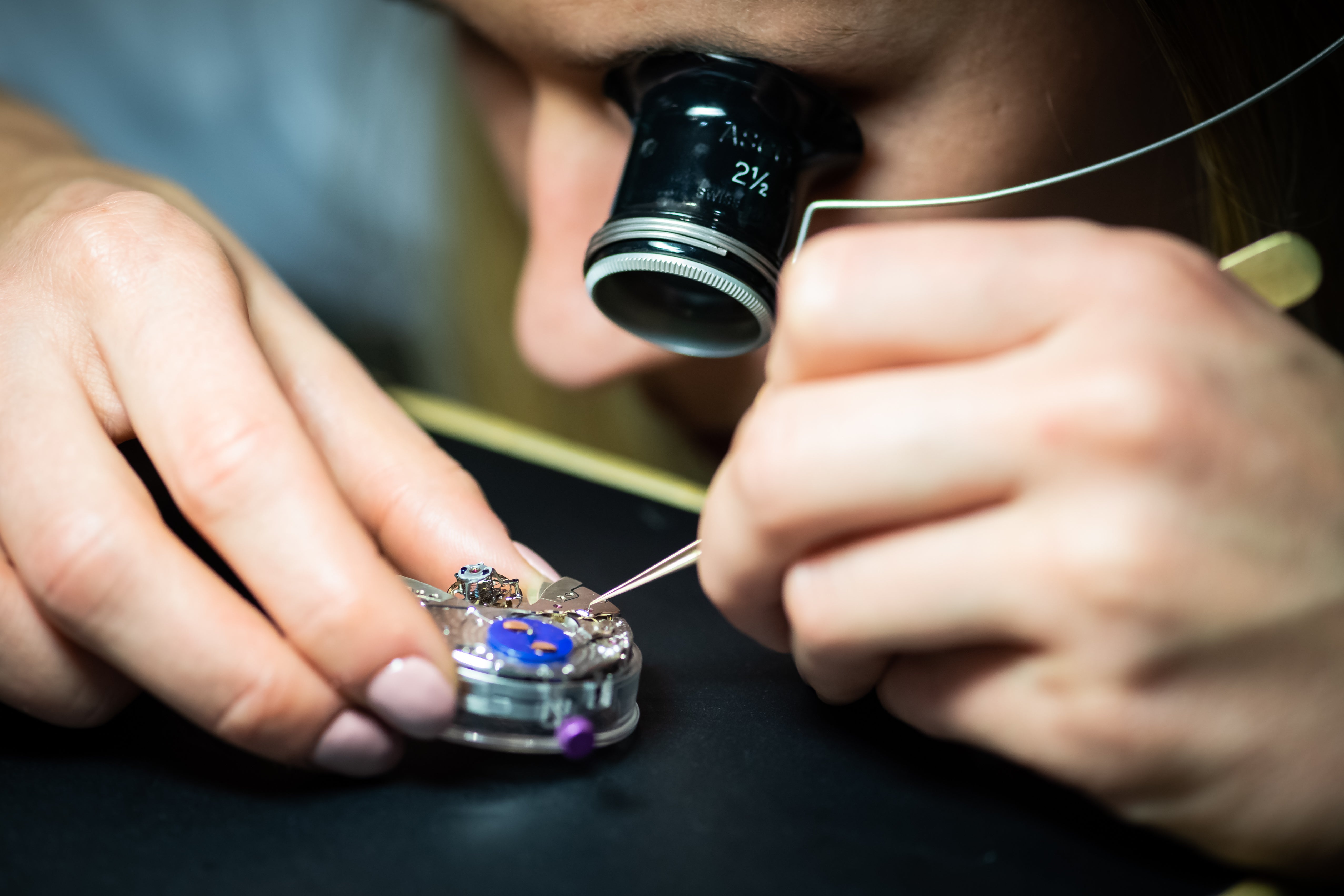 Homme qui porte une loupe et qui manipule un mouvement de montre à l'aide d'une brucelle.