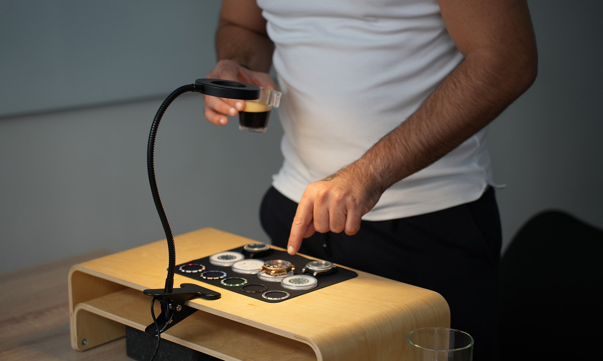 Un homme assemble une montre lors d’un atelier d’horlogerie. Il tient un café dans une main et pointe du doigt des cadrans de montre disposés sur un plateau en bois, éclairés par une lampe flexible.