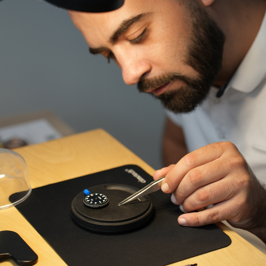 Un homme assemble une montre lors d’un atelier d’horlogerie. Il tient un outil horloger dans la main, une brucelle, et attrape une aiguille afin de la positionner sur le cadran de la montre.